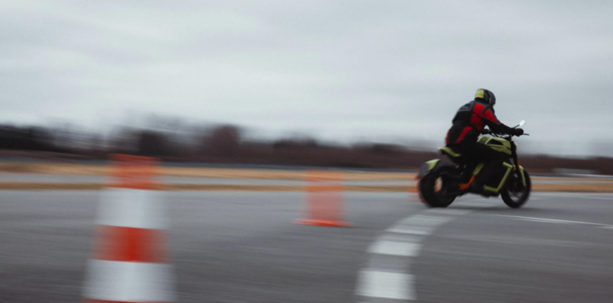 A motorcyclist wearing a black and red outfit rides at high speed around a curve on a track, with orange traffic cones marking the path. The background is blurred, indicating motion.