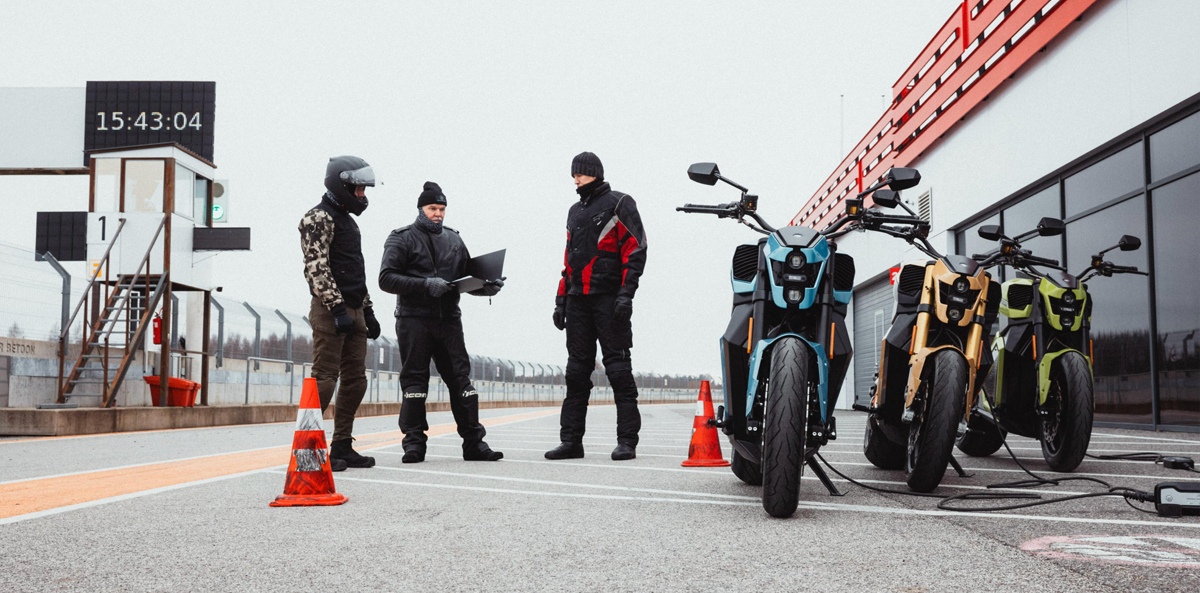 Three people in motorcycle gear stand on a racetrack near three electric motorcycles that are charging. Two traffic cones are nearby, and a clock shows 15:43:04. Overcast sky and a track building are in the background.