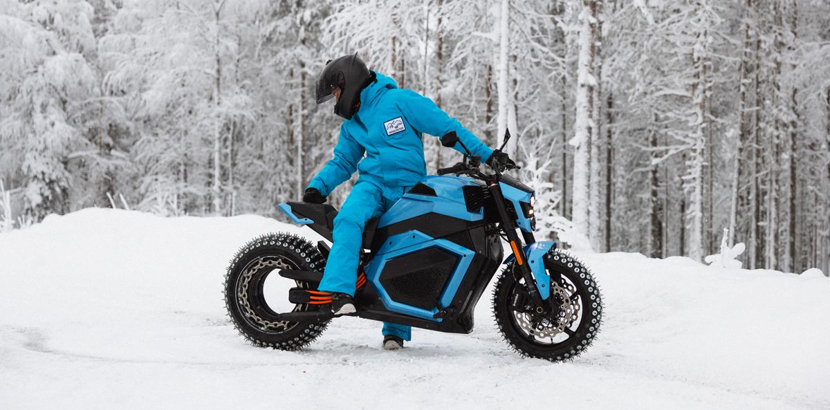 A person wearing a blue snowsuit and black helmet sits on a blue electric motorcycle with studded tires, surrounded by a snowy landscape with frosted trees in the background.