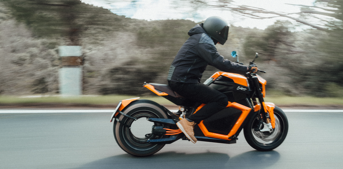 A person wearing a helmet rides a bright orange motorcycle on a road, with a blurred background of trees and sky, suggesting high speed. The rider is dressed in dark clothing and the road appears smooth and winding.
