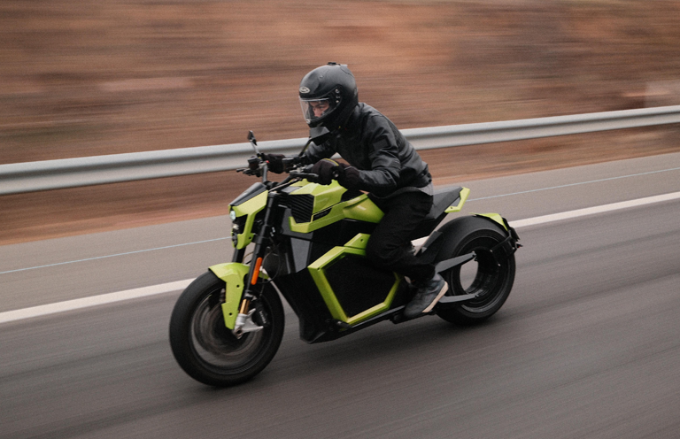 A person wearing a black helmet and jacket rides a bright green motorcycle at speed on a road. The background appears blurred, indicating motion. The road is bordered by a metal guardrail.