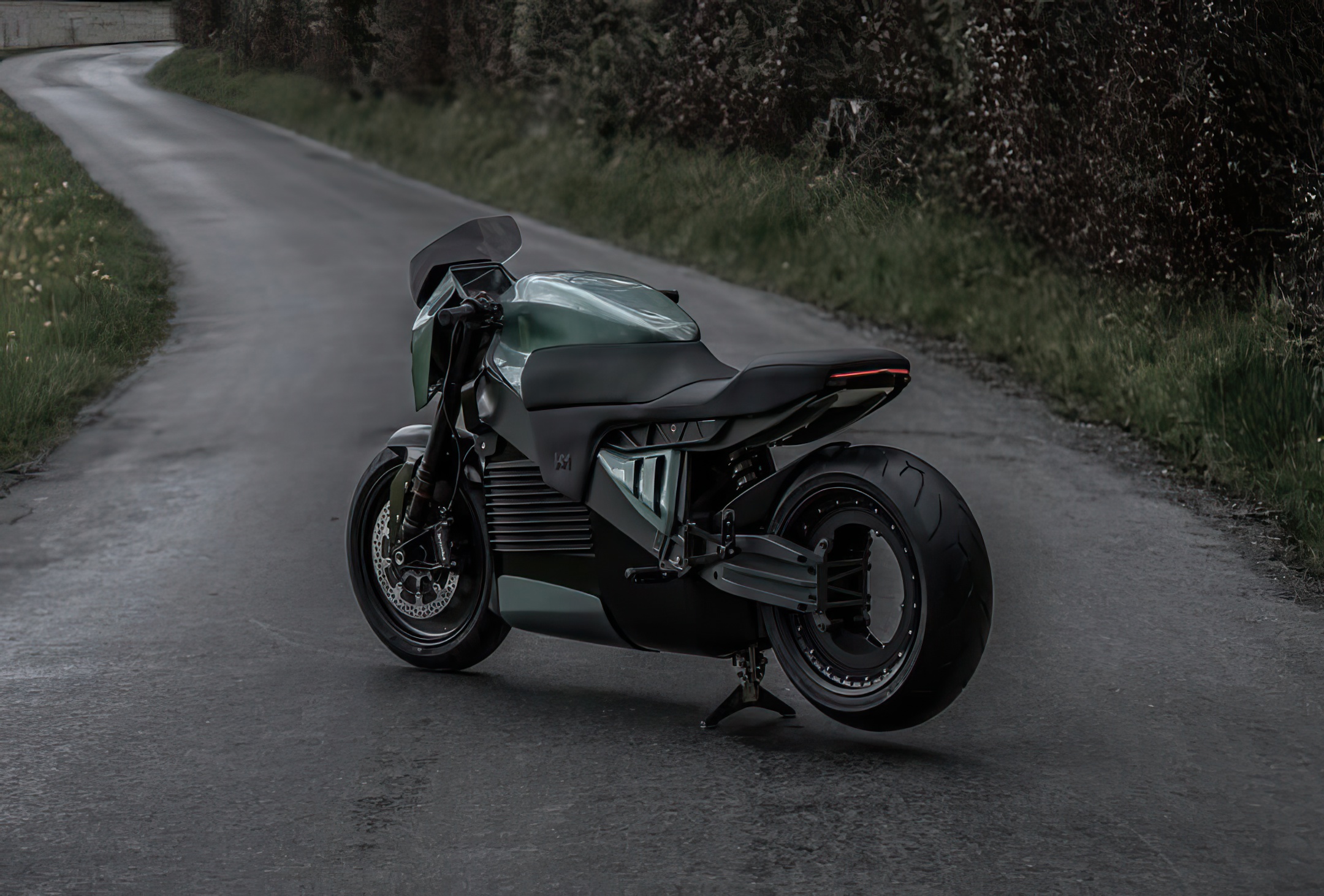A sleek, modern motorcycle with a matte black and metallic green finish is parked on an empty, winding rural road bordered by grass and trees under an overcast sky.